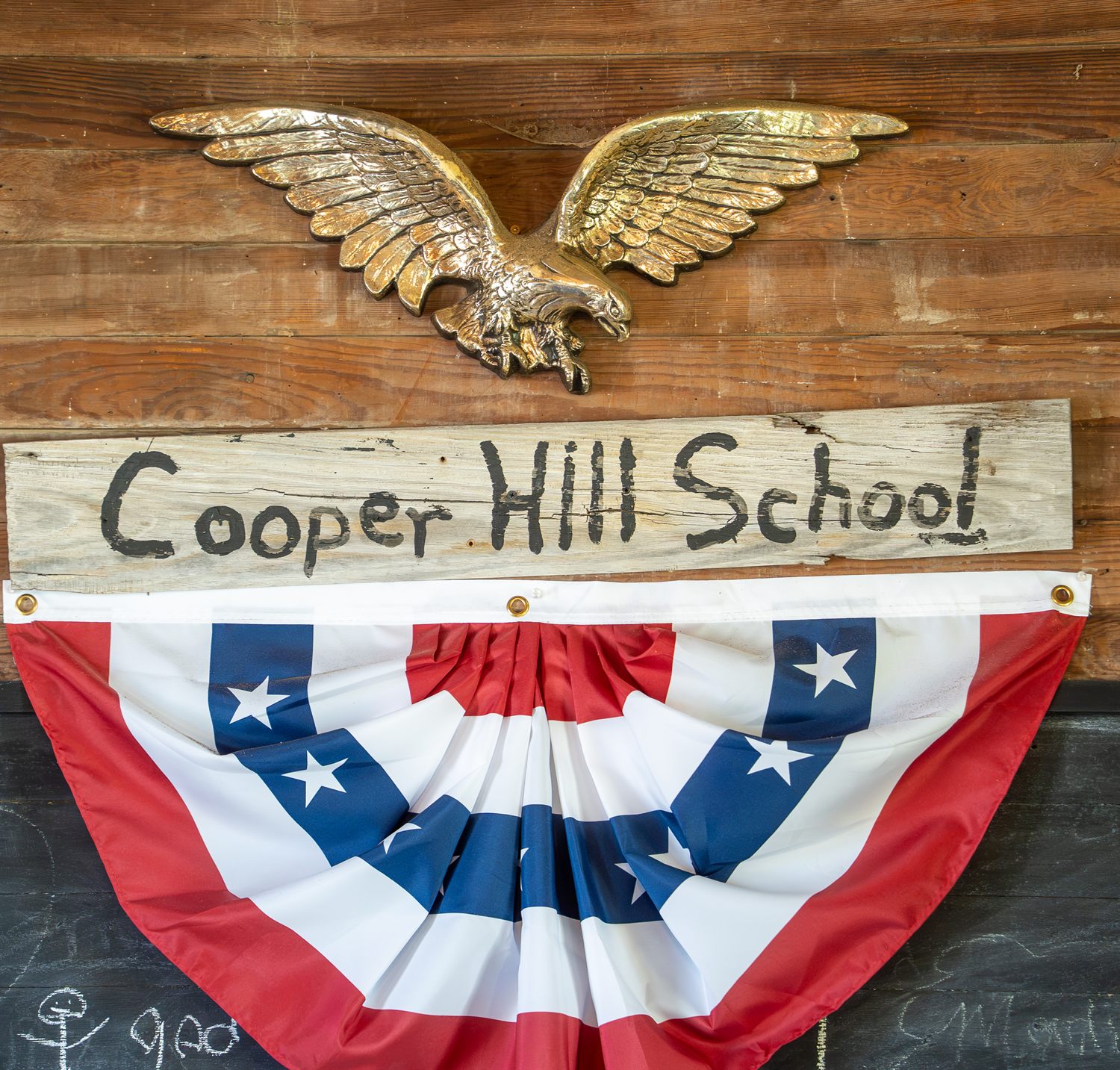 The original hand-painted Cooper Hill School sign hanging on a weathered plank wall beneath a brass eagle and a patriotic bunting.